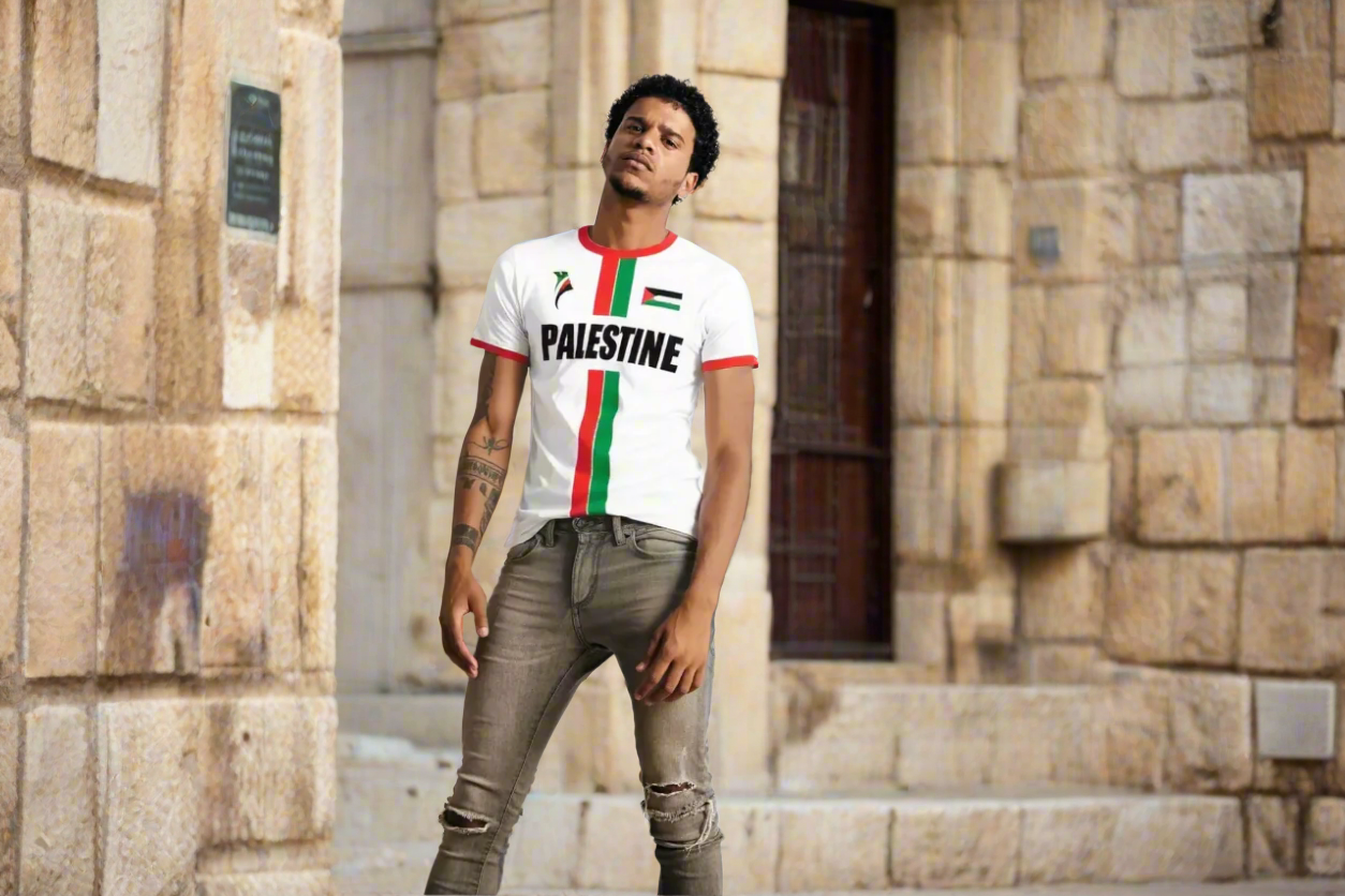 Person wearing a white t-shirt with 'PALESTINE' text and red, green, and white stripes in front of a stone building .