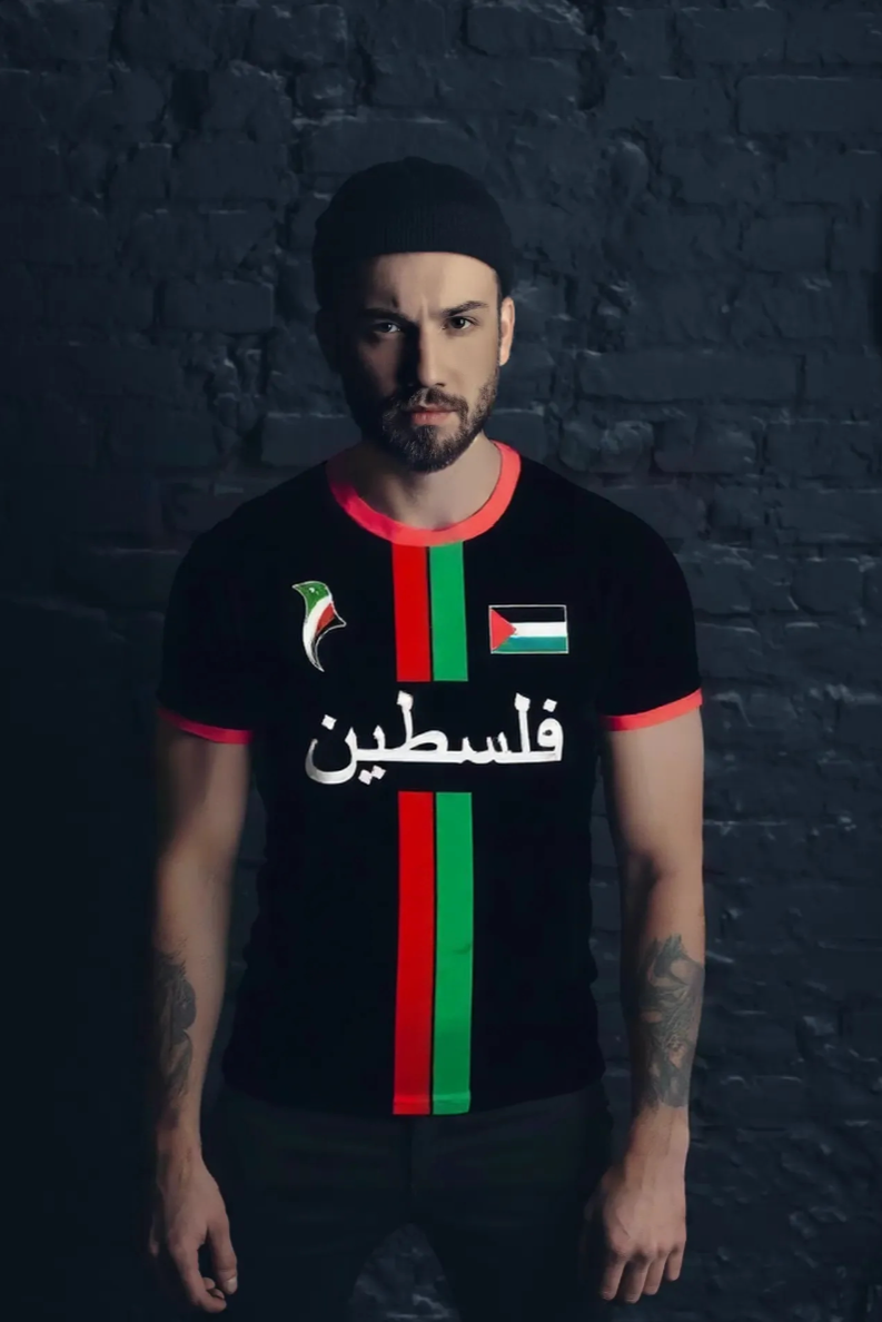 Person wearing a black t-shirt with red and green stripes and Arabic text palestine against a dark brick wall.