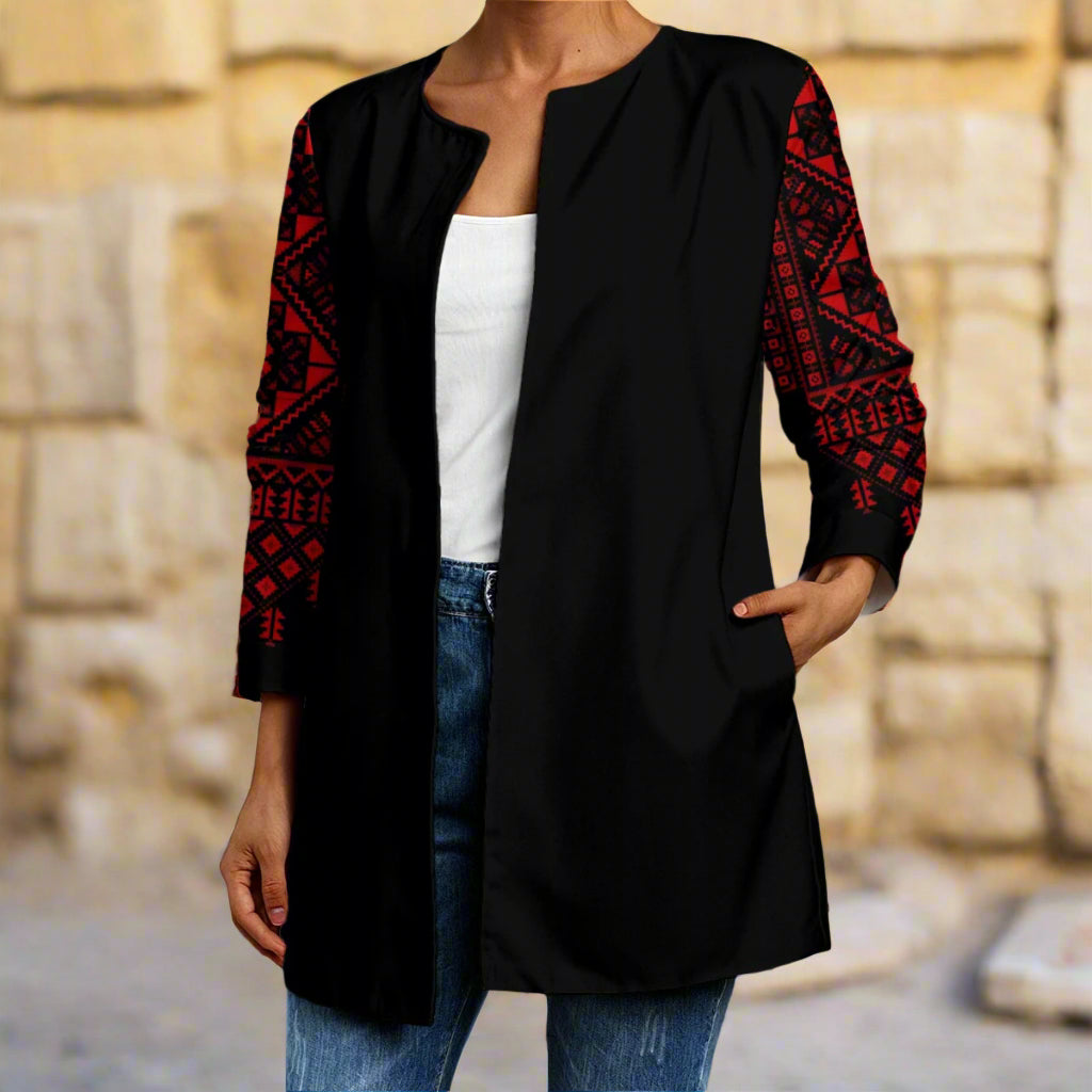 Person wearing a black cardigan with red patterns of tatreez palestinian print, white shirt, and blue jeans in front of a stone wall.