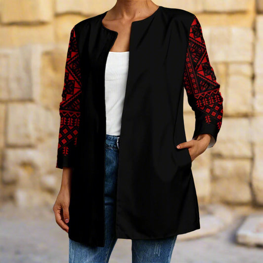 Person wearing a black cardigan with red patterns of tatreez palestinian print, white shirt, and blue jeans in front of a stone wall.