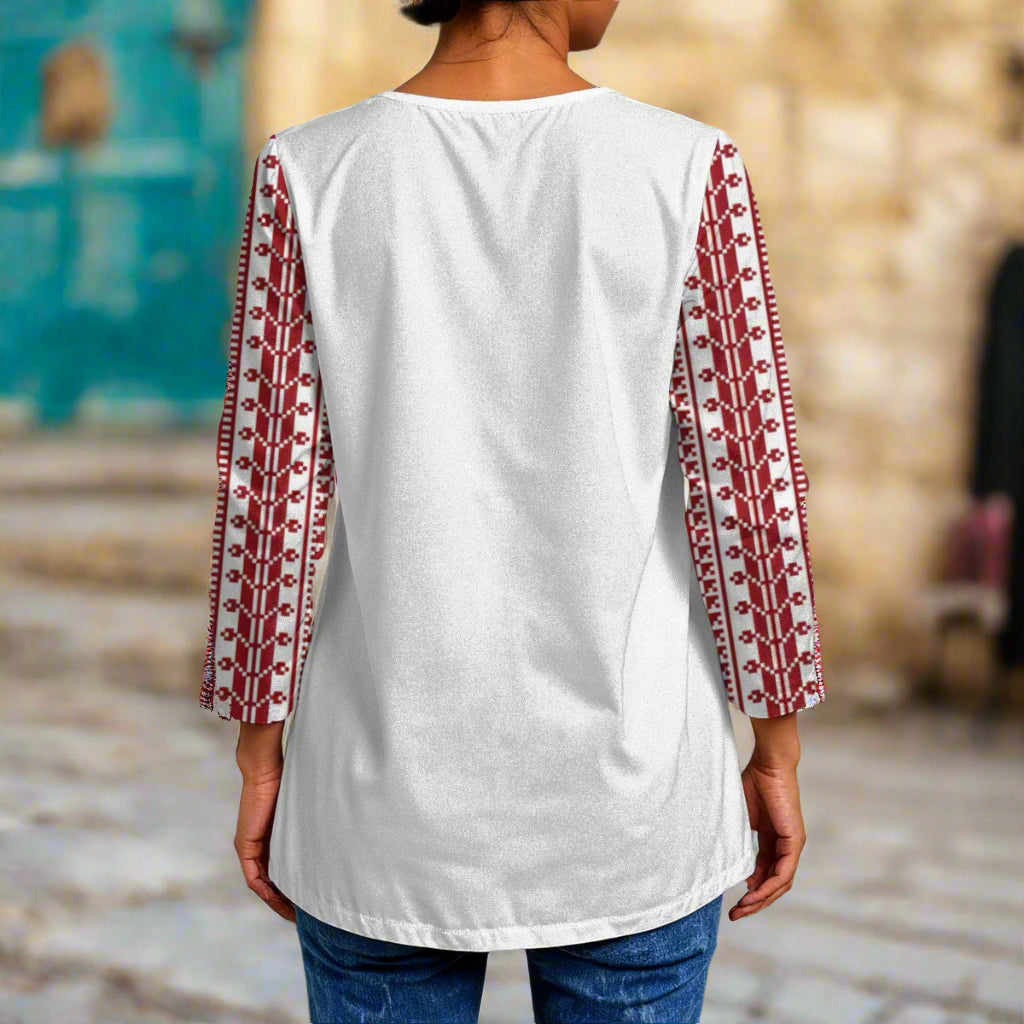 Person wearing a white top with red patterned tatreez print palestinian sleeves in an outdoor setting.