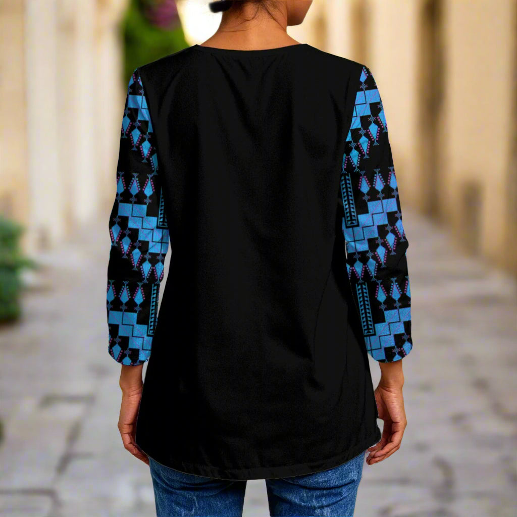 Person wearing a black top with blue patterned sleeves with tatreez blue palestinian pattern standing in front of a textured wall.