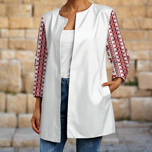 Person wearing a white cardigan with red tatreez print embroidery from palestine standing against a stone wall.
